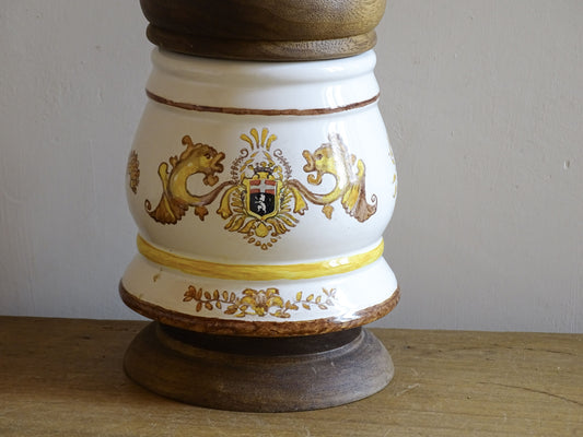A vintage ceramic apothecary jar with hand-painted decoration and gilt lettering, with a wooden lid and base. Inspired by traditional European pharmacy vessels.