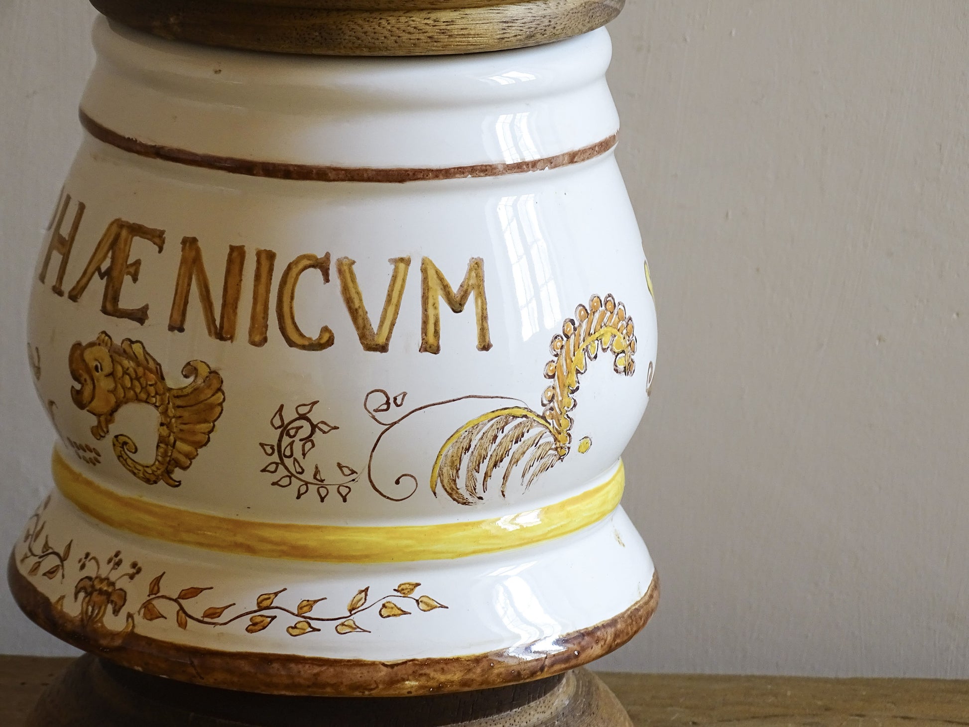 A vintage ceramic apothecary jar with hand-painted decoration and gilt lettering, with a wooden lid and base. Inspired by traditional European pharmacy vessels.