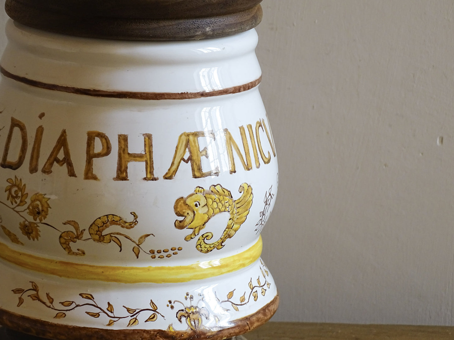 A vintage ceramic apothecary jar with hand-painted decoration and gilt lettering, with a wooden lid and base. Inspired by traditional European pharmacy vessels.