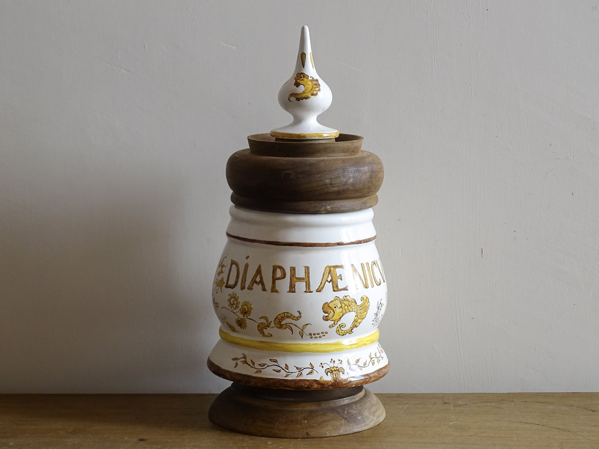 A vintage ceramic apothecary jar with hand-painted decoration and gilt lettering, with a wooden lid and base. Inspired by traditional European pharmacy vessels.