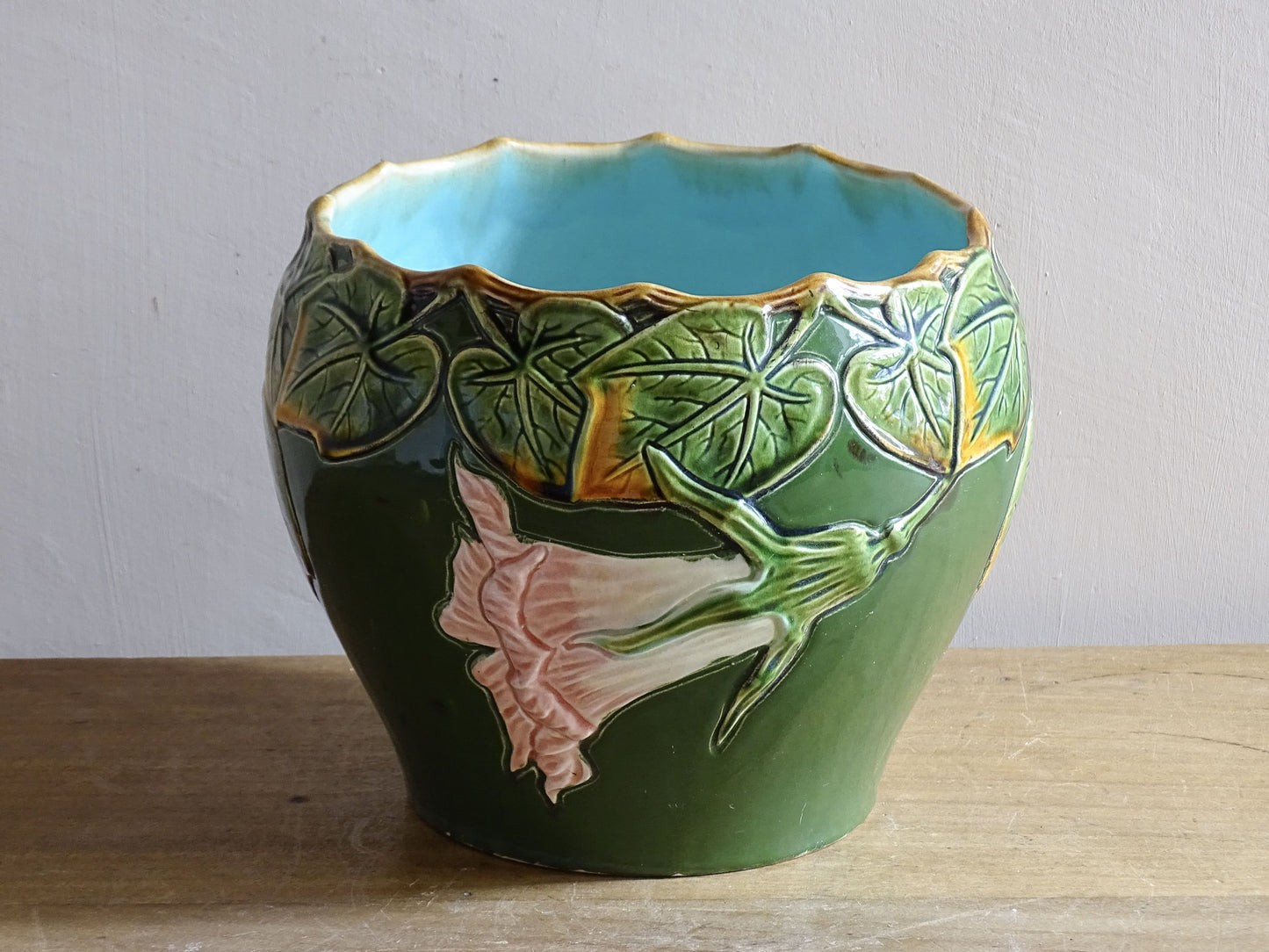 An antique ceramic cache pot dating to the turn of the 20th century, with strong Art Nouveau influence. The planter is richly decorated in relief, with trailing ivy leaves encircling the rim and a flowing trumpet-shaped flower applied to the body.
