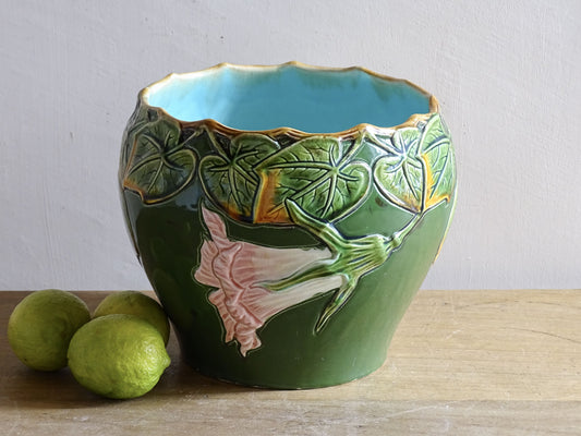 An antique ceramic cache pot dating to the turn of the 20th century, with strong Art Nouveau influence. The planter is richly decorated in relief, with trailing ivy leaves encircling the rim and a flowing trumpet-shaped flower applied to the body.
