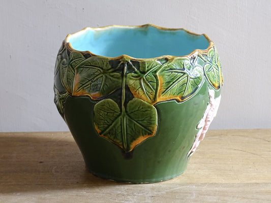 An antique ceramic cache pot dating to the turn of the 20th century, with strong Art Nouveau influence. The planter is richly decorated in relief, with trailing ivy leaves encircling the rim and a flowing trumpet-shaped flower applied to the body.