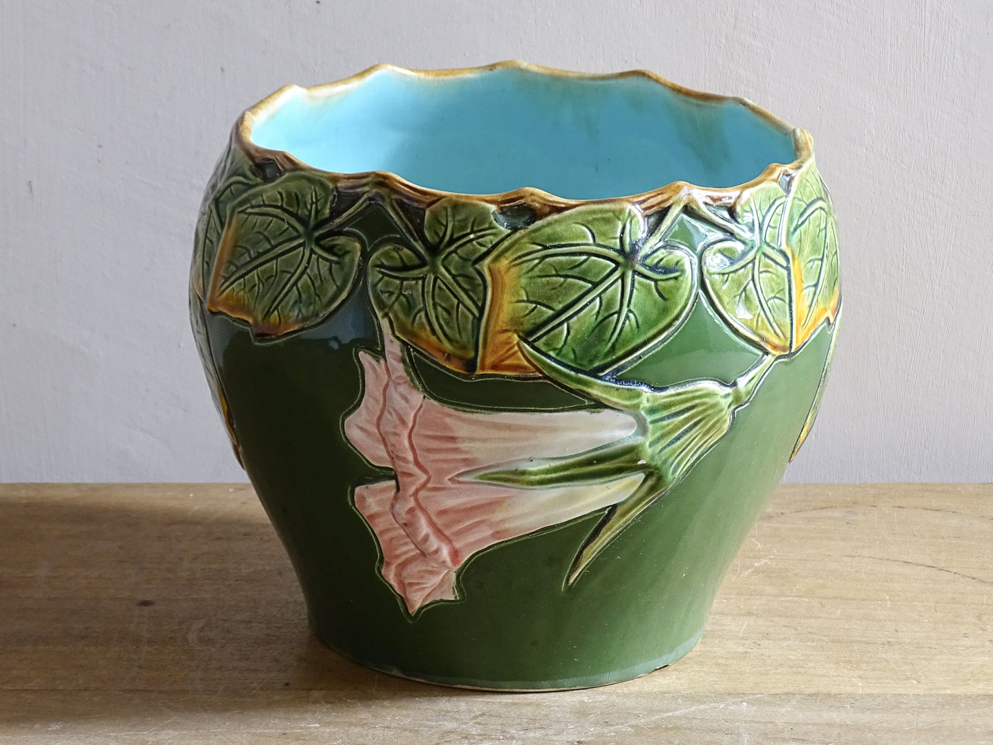 An antique ceramic cache pot dating to the turn of the 20th century, with strong Art Nouveau influence. The planter is richly decorated in relief, with trailing ivy leaves encircling the rim and a flowing trumpet-shaped flower applied to the body.