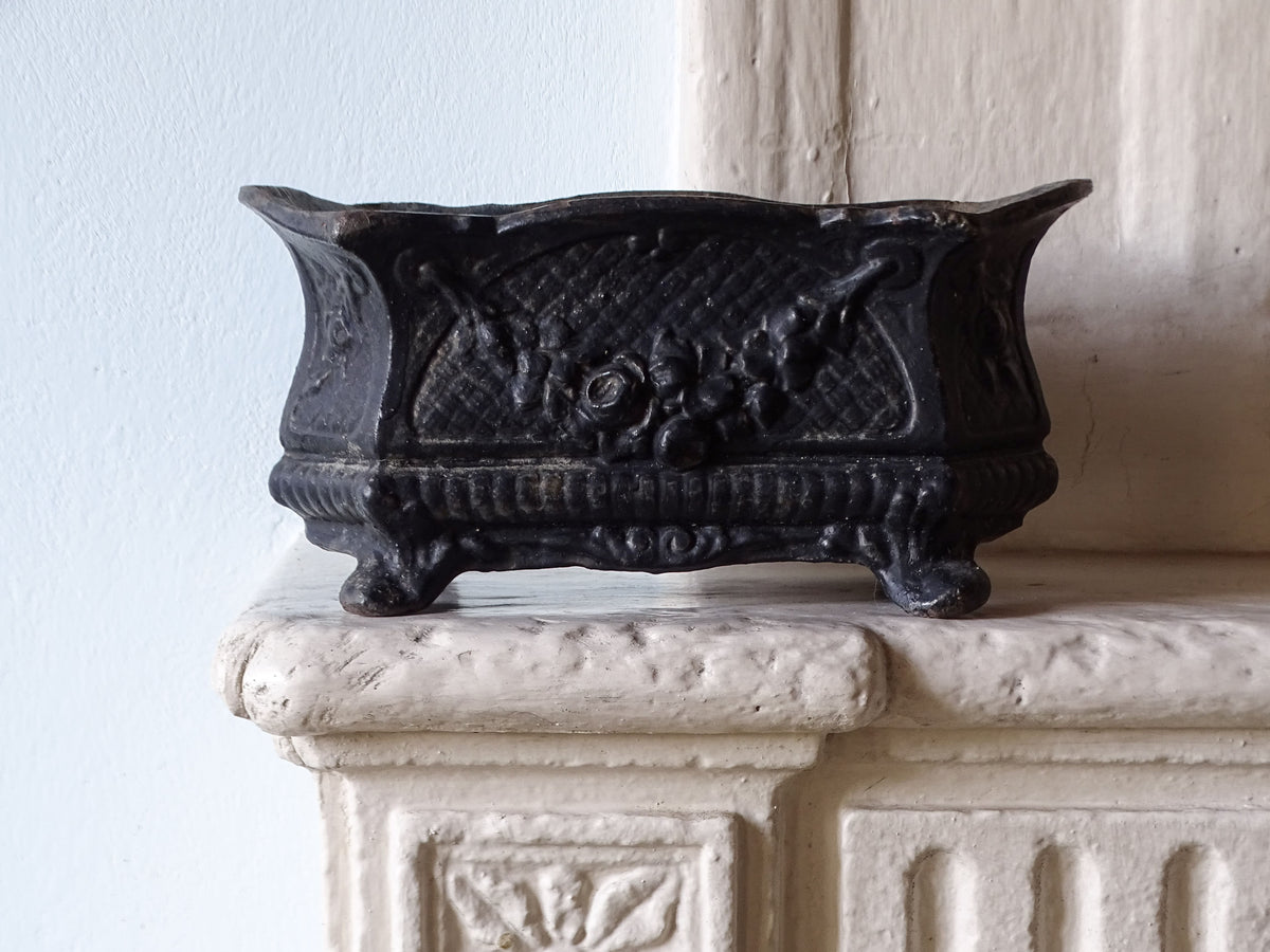 French antique black cast iron planter with ornate floral carvings and feet, displayed on a stone mantelpiece against a pale blue wall.