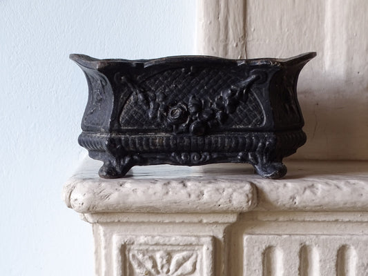 French antique black cast iron planter with ornate floral carvings and feet, displayed on a stone mantelpiece against a pale blue wall.