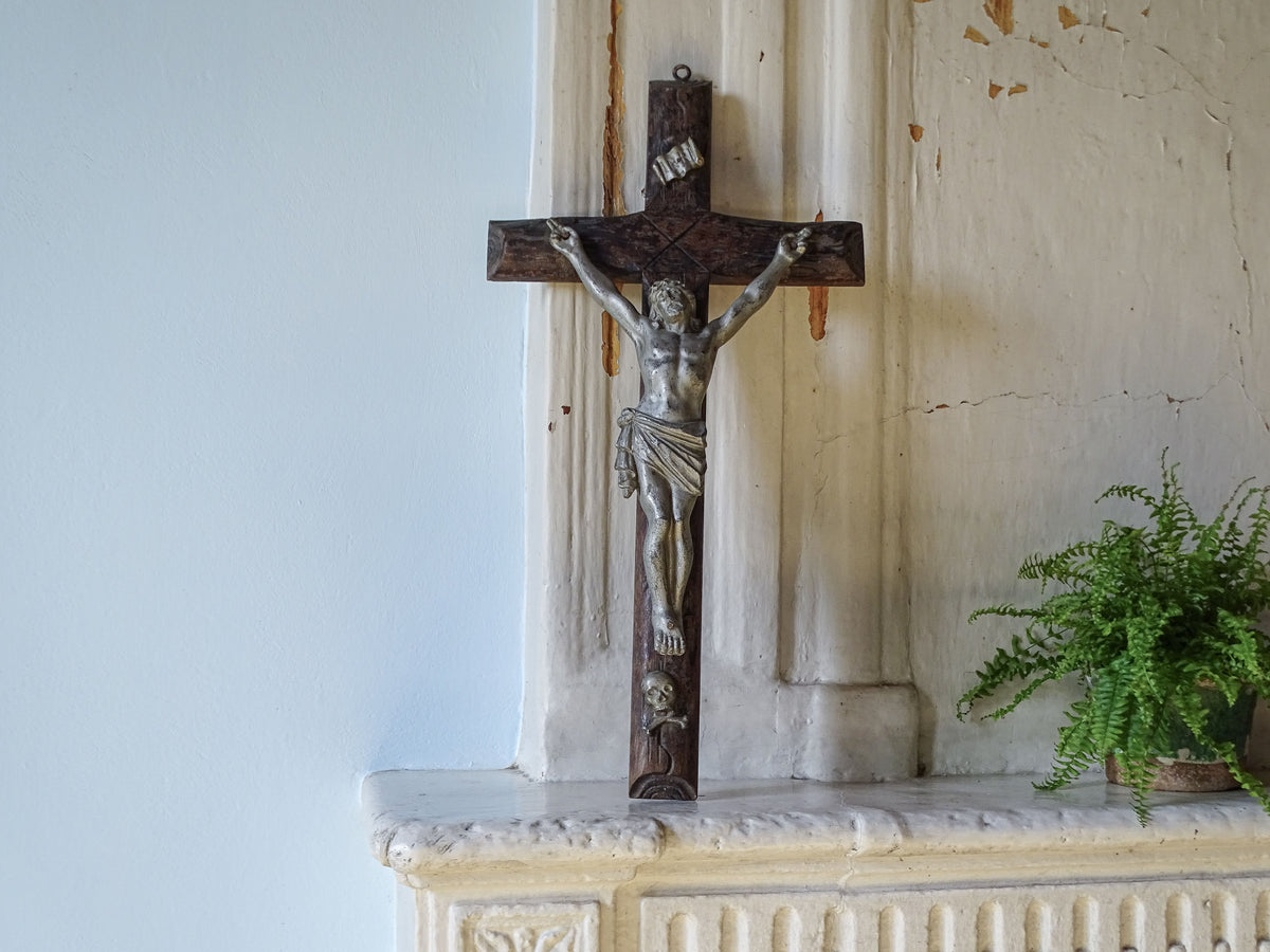 A dark wooden French antique crucifix with a silver-toned metal corpus of Jesus Christ, displayed on a white stone mantelpiece against a light blue wall.