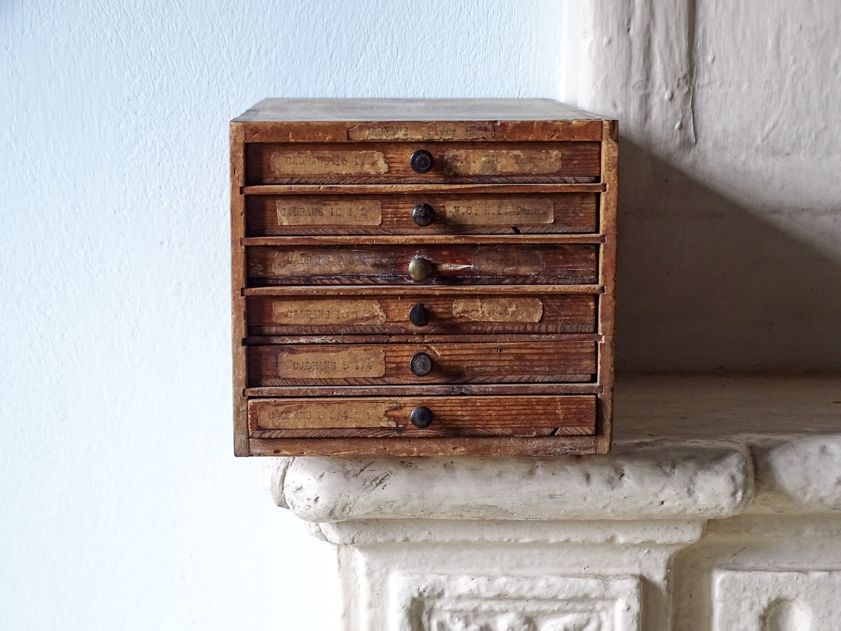 French antique wooden watchmaker's cabinet with five small drawers and dark round knobs, displayed on a white stone mantelpiece against a light blue wall.