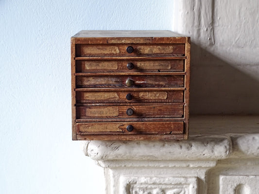 French antique wooden watchmaker's cabinet with five small drawers and dark round knobs, displayed on a white stone mantelpiece against a light blue wall.
