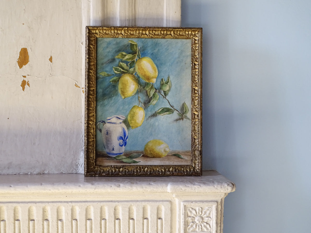 A framed still life painting of lemons on a branch with a blue-and-white ceramic pitcher, displayed on a white fireplace mantel against a pale blue wall.