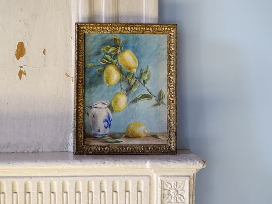 A framed still life painting of lemons on a branch with a blue-and-white ceramic pitcher, displayed on a white fireplace mantel against a pale blue wall.