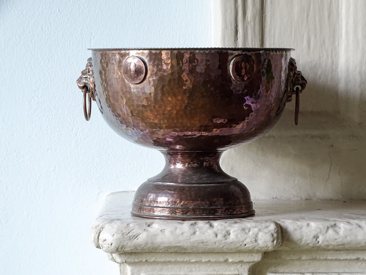 French antique hammered copper champagne bucket with lion head handles, pictured on a country house fireplace next to a pale blue wall. 