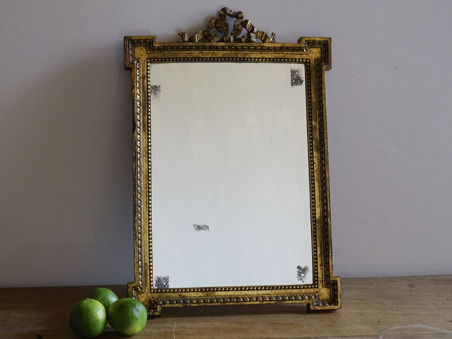 French vintage gilt mirror with ribbon crest and foxed glass displayed on a wooden surface