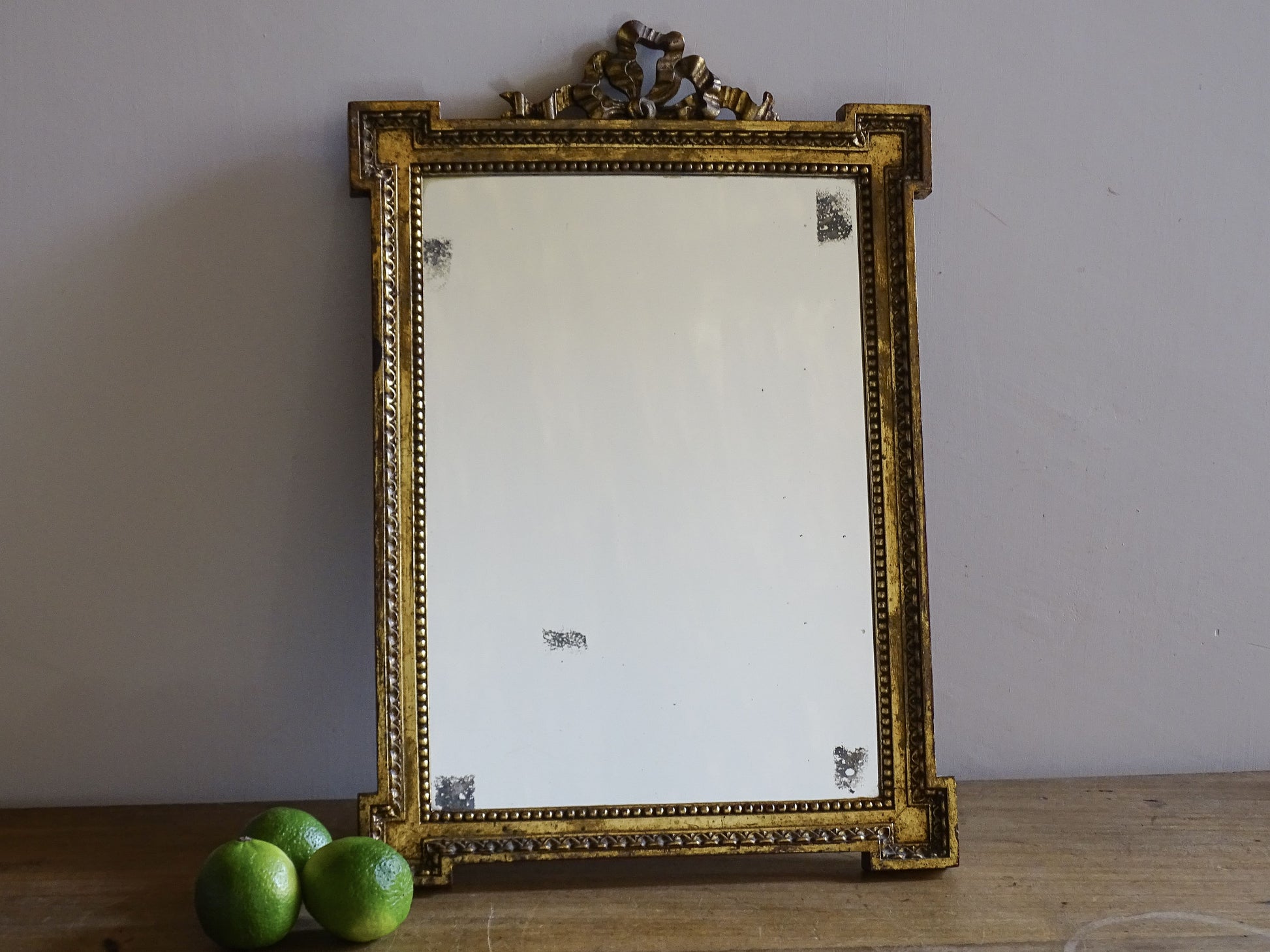 French vintage gilt mirror with ribbon crest and foxed glass displayed on a wooden surface