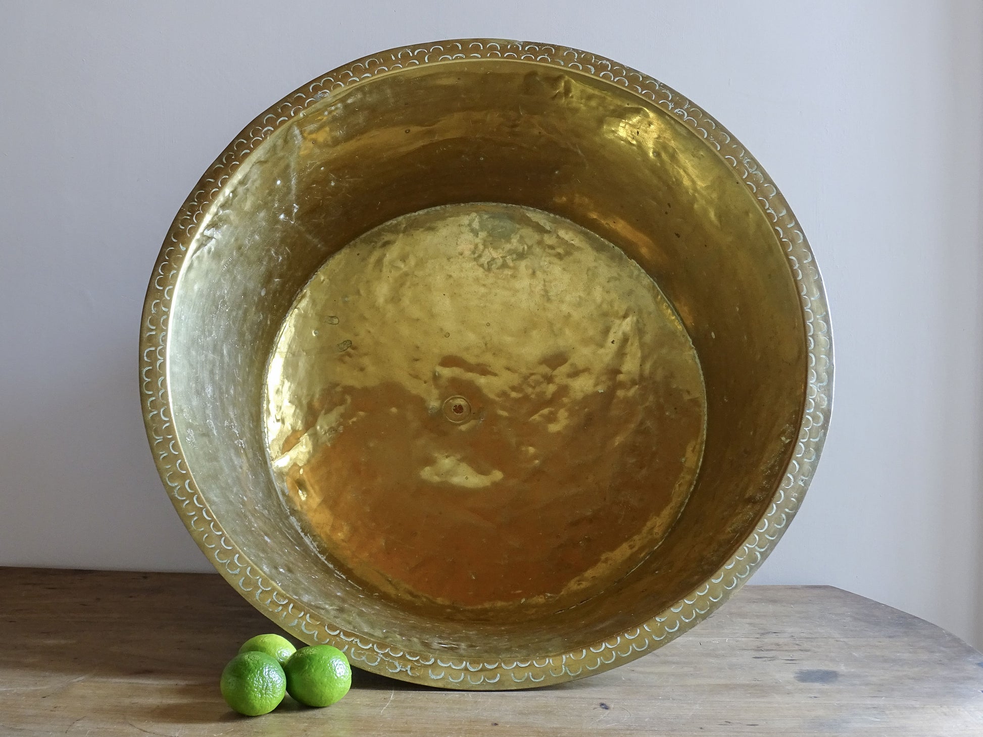 Large antique brass basin displayed upright with limes for scale, showing its golden interior and decorated rim.