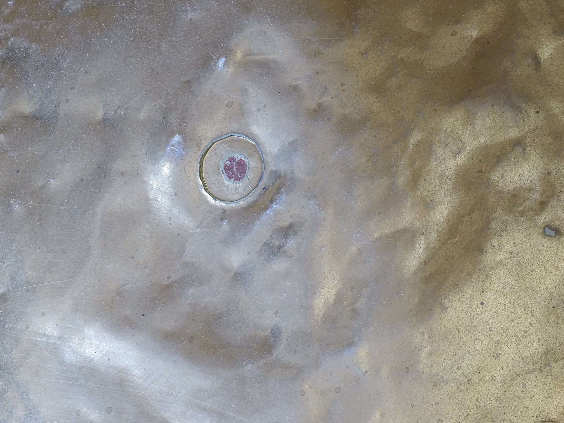 Circular metal plug at the centre of an antique brass basin, possibly sealing an original drainage opening.
