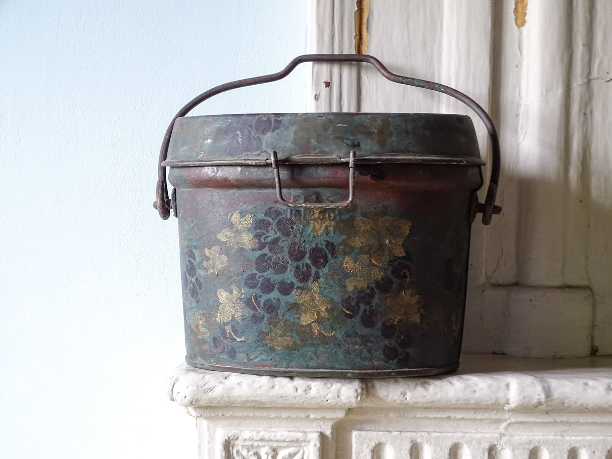 Vintage toleware box with handle and lid, decorated with painted grapes and vines in purple and gold, displayed on a country house fireplace