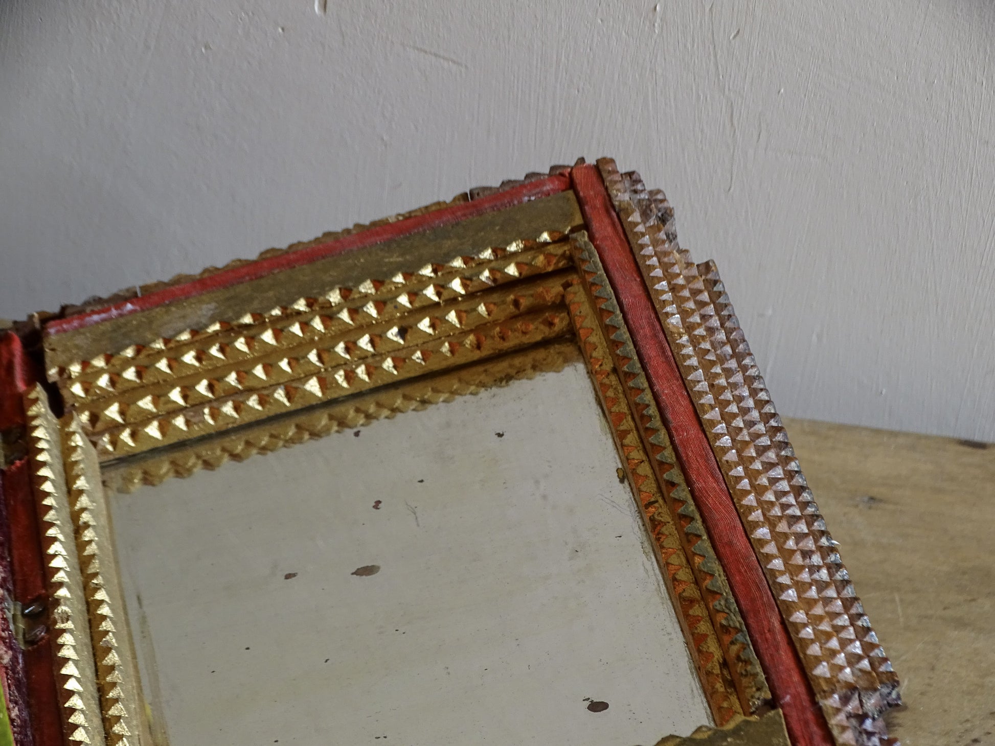 Close-up of the aged mirror inside the lid of an antique tramp art box, surrounded by gilt carved wood and narrow red trim.