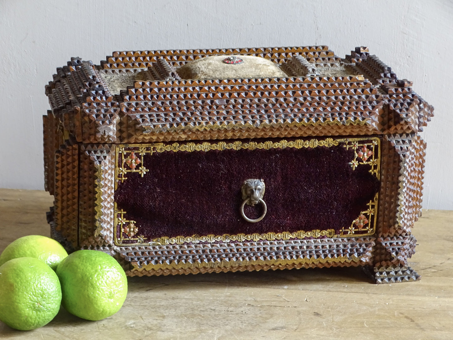 Front view of ornate antique tramp art box with burgundy velvet panel, lion-mask ring handle and raised fabric pincushion.