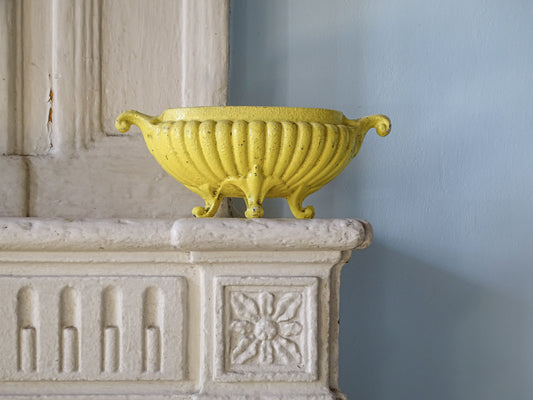 Antique French cast iron jardinière planter with worn yellow painted finish and scrolling handles displayed on a country fireplace