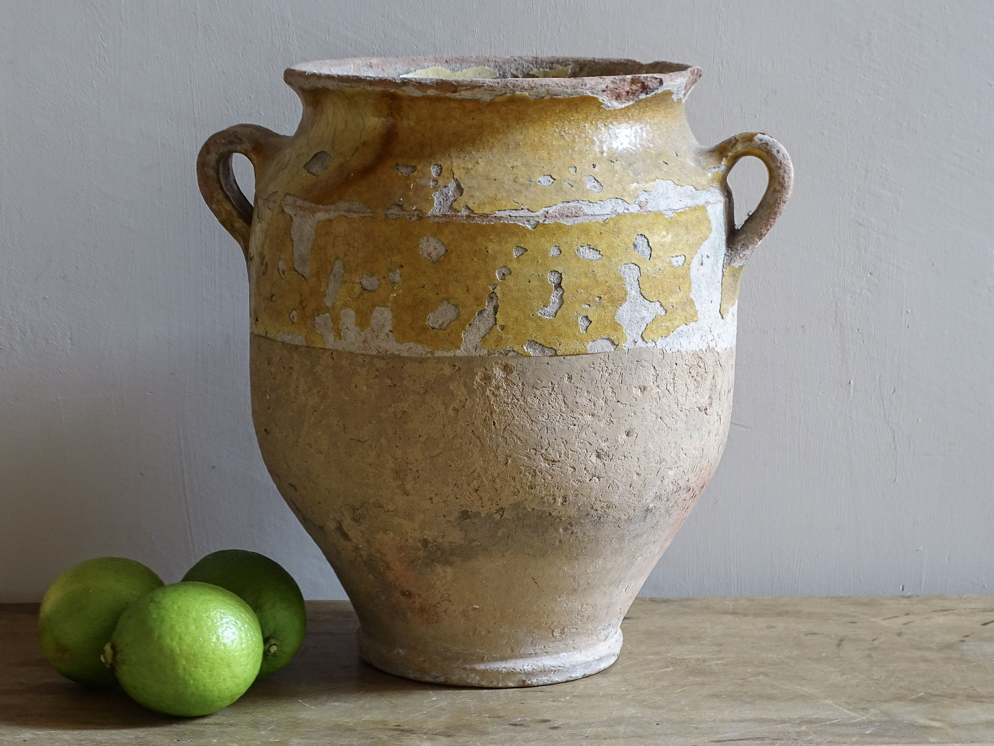 19th-century French confit pot with worn mustard-yellow glaze, twin handles and three green limes for scale.