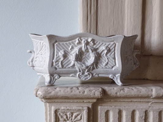 French antique cast iron planter in white with four feet and ornate detailing, pictured on a country house fireplace. 