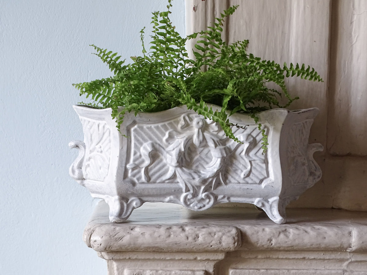 An ornate French antique white cast-iron planter with carved floral details and small feet, filled with green ferns, displayed on a stone mantelpiece against a light blue wall.