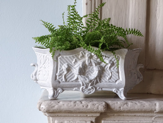 An ornate French antique white cast-iron planter with carved floral details and small feet, filled with green ferns, displayed on a stone mantelpiece against a light blue wall.