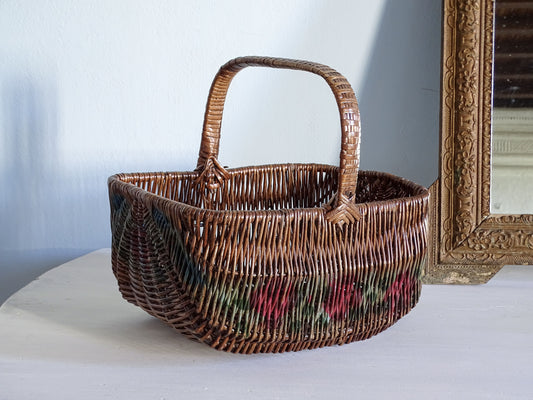 A French vintage woven wicker basket with a floral pattern, displayed on a white table next to an ornate gold mirror frame.