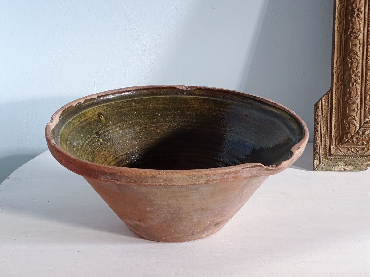 Rustic French antique tian bowl with olive green glaze interior, pictured in front of a pale blue wall