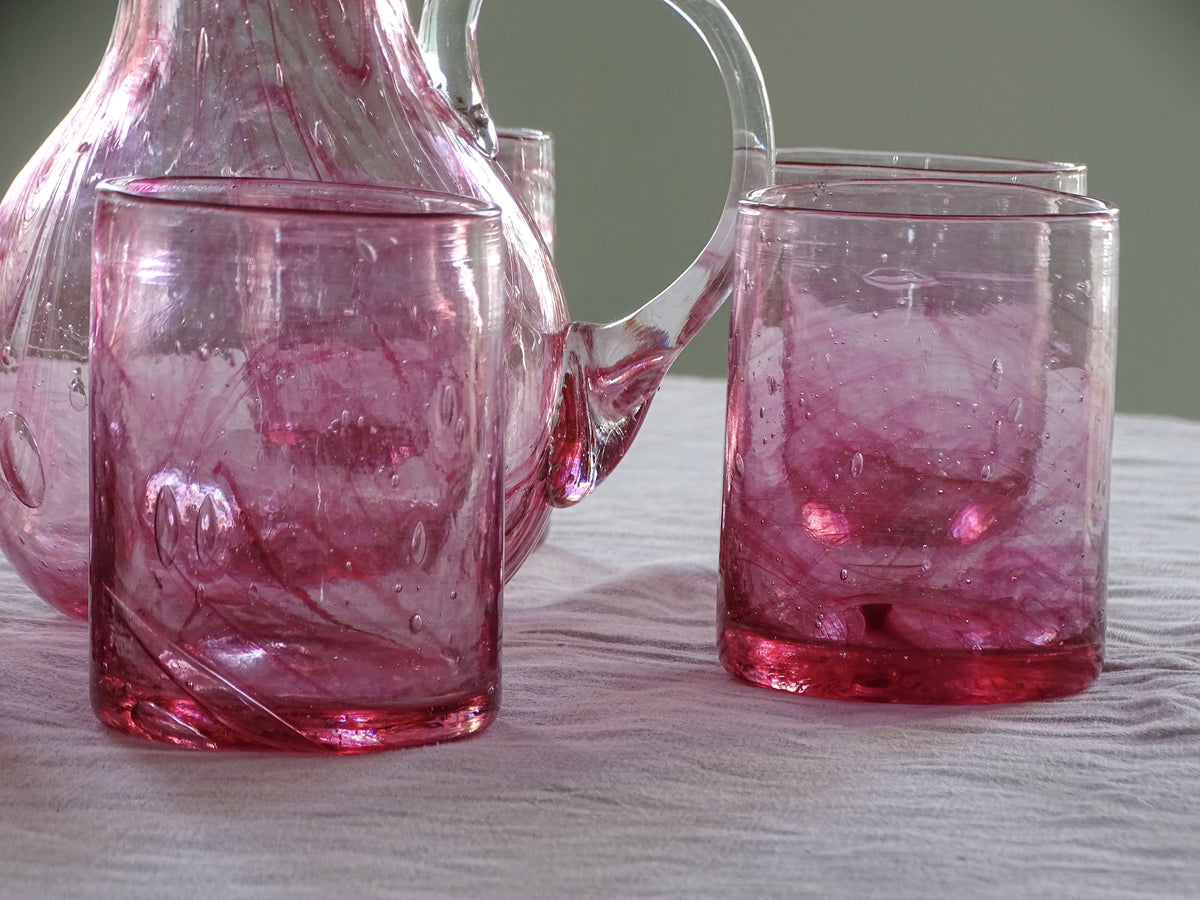 A pink tinted glass carafe and matching tumblers with a swirled, textured pattern, arranged on a light-colored tablecloth.