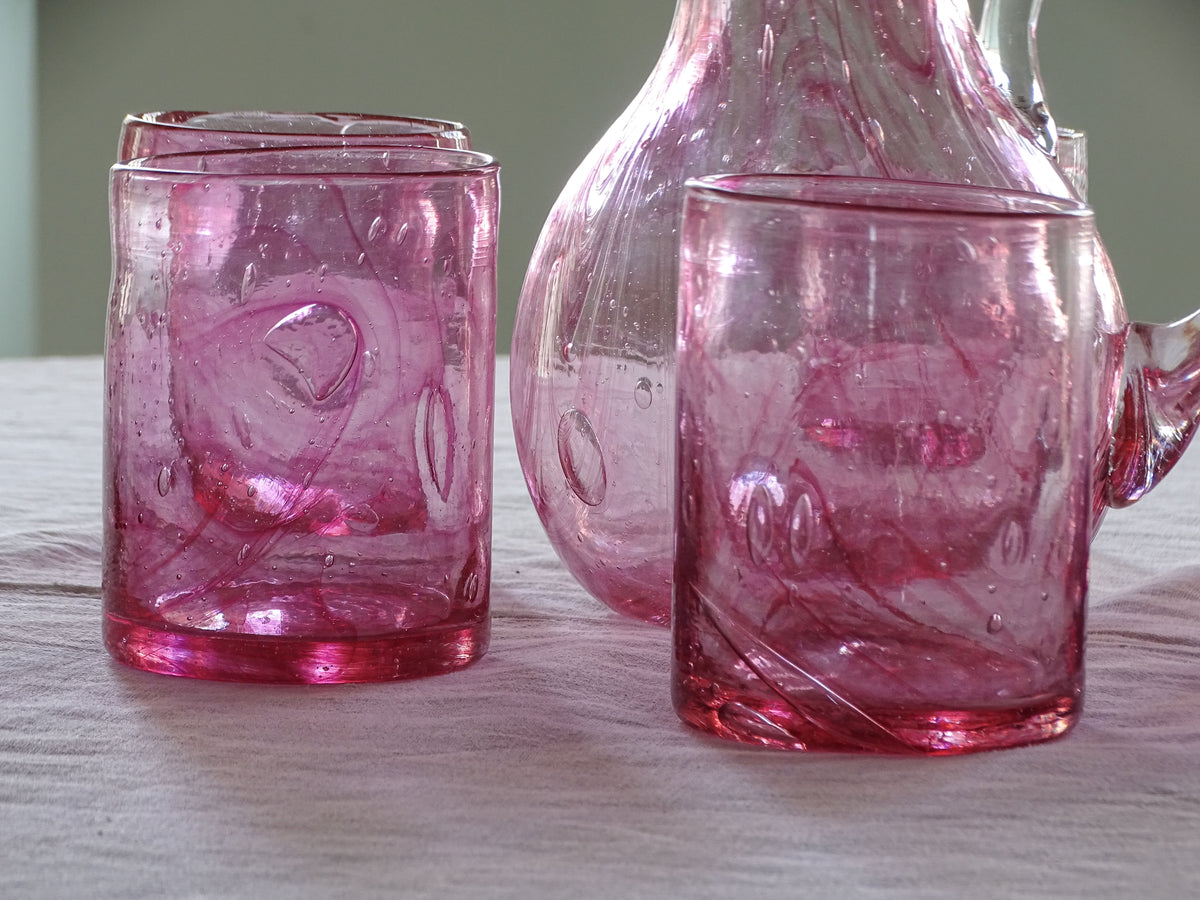 A set of french vintage pink glass tableware, including two textured tumblers and a matching glass jug, arranged on a light-colored tablecloth.