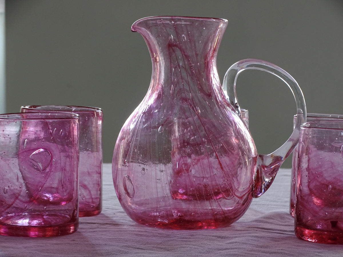 French vintage pink glass pitcher with a curved handle and matching pink drinking glasses arranged on a table. 