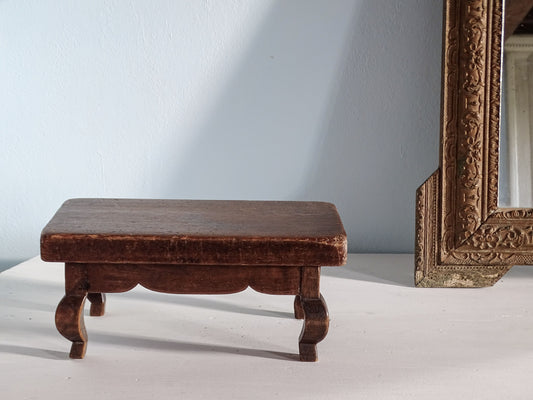 A small dark brown wooden stool with curved legs, shown on a white surface against a pale blue wall, with part of an ornate gold-framed mirror visible on the right.