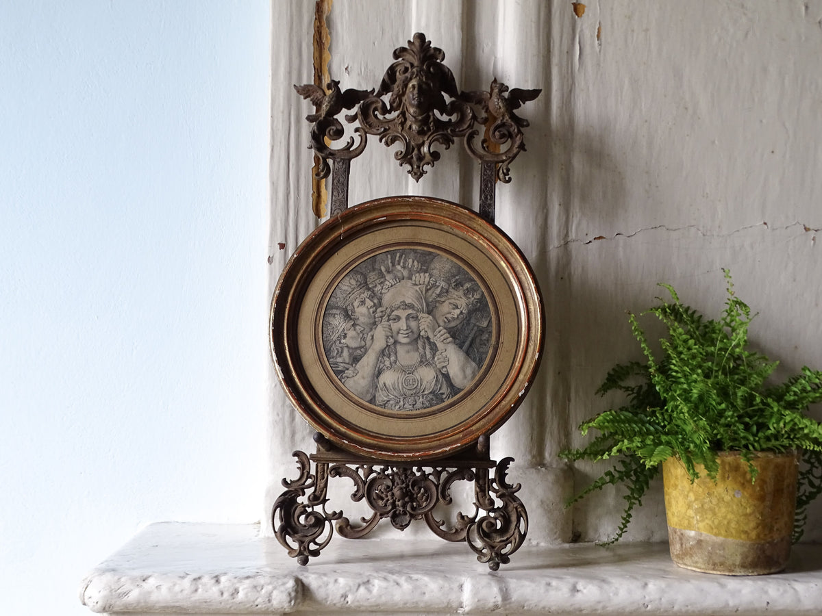 Antique German cast iron tabletop easel displaying a circular framed engraving on a stone mantelpiece beside a potted fern