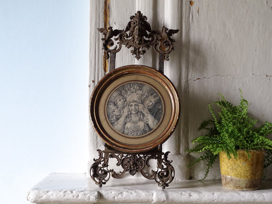 Antique German cast iron tabletop easel displaying a circular framed engraving on a stone mantelpiece beside a potted fern