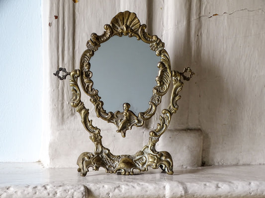 French vintage brass dressing table mirror in Rococo style, with an ornate freestanding base, cherub figures and scallop shell crest