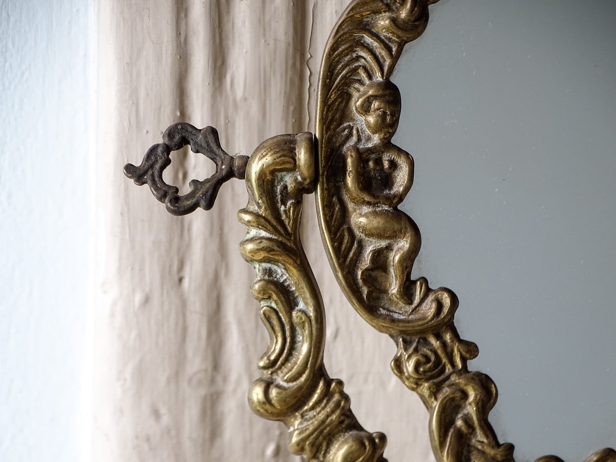 Ornate brass mirror frame featuring a sculpted cherub, scrolling foliage and decorative key-shaped adjustment screw