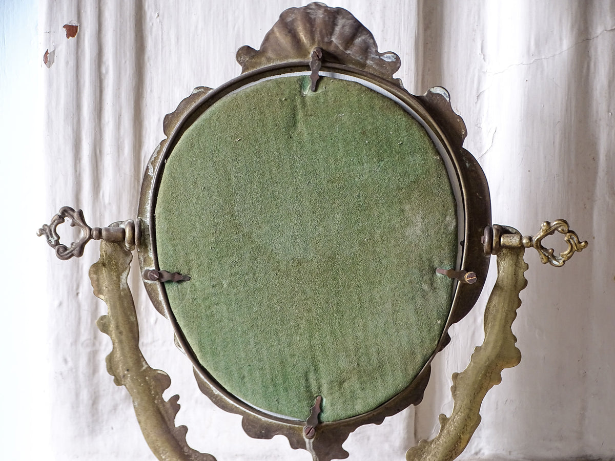 Side view of a freestanding vintage brass vanity mirror showing the adjustable oval frame and ornate supporting base