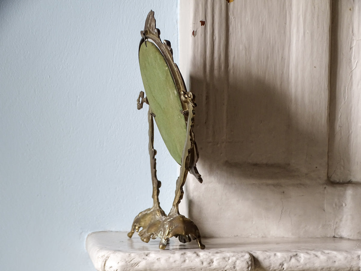 Rear of a vintage brass dressing table mirror showing the green fabric backing, metal retaining clips and adjustable side screws