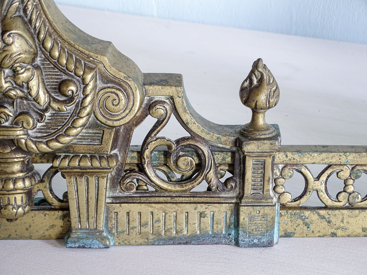 Detail of brass fireplace fender with scrolling foliage, fluted architectural supports and flame finial