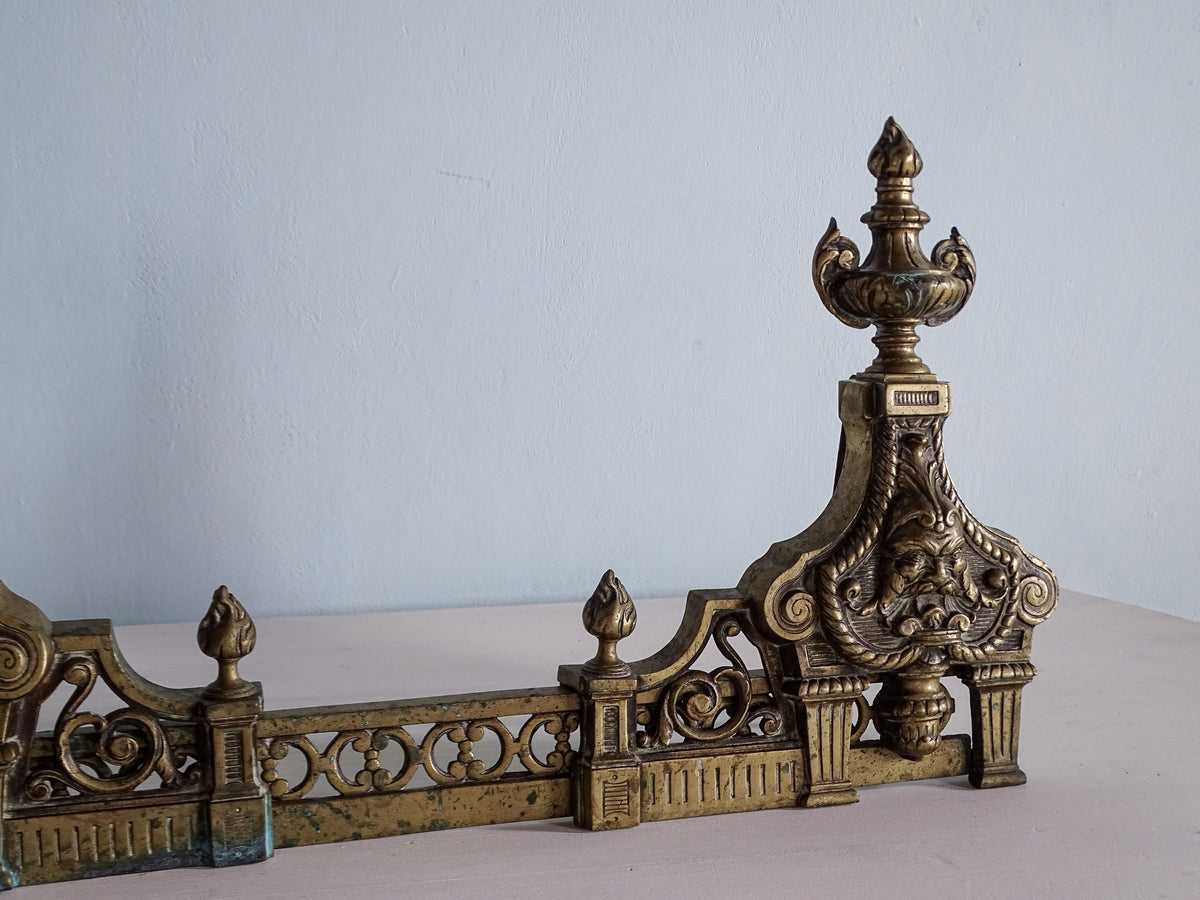Right upright of antique French brass fireplace fender showing ornate mask, urn finial and connecting rail