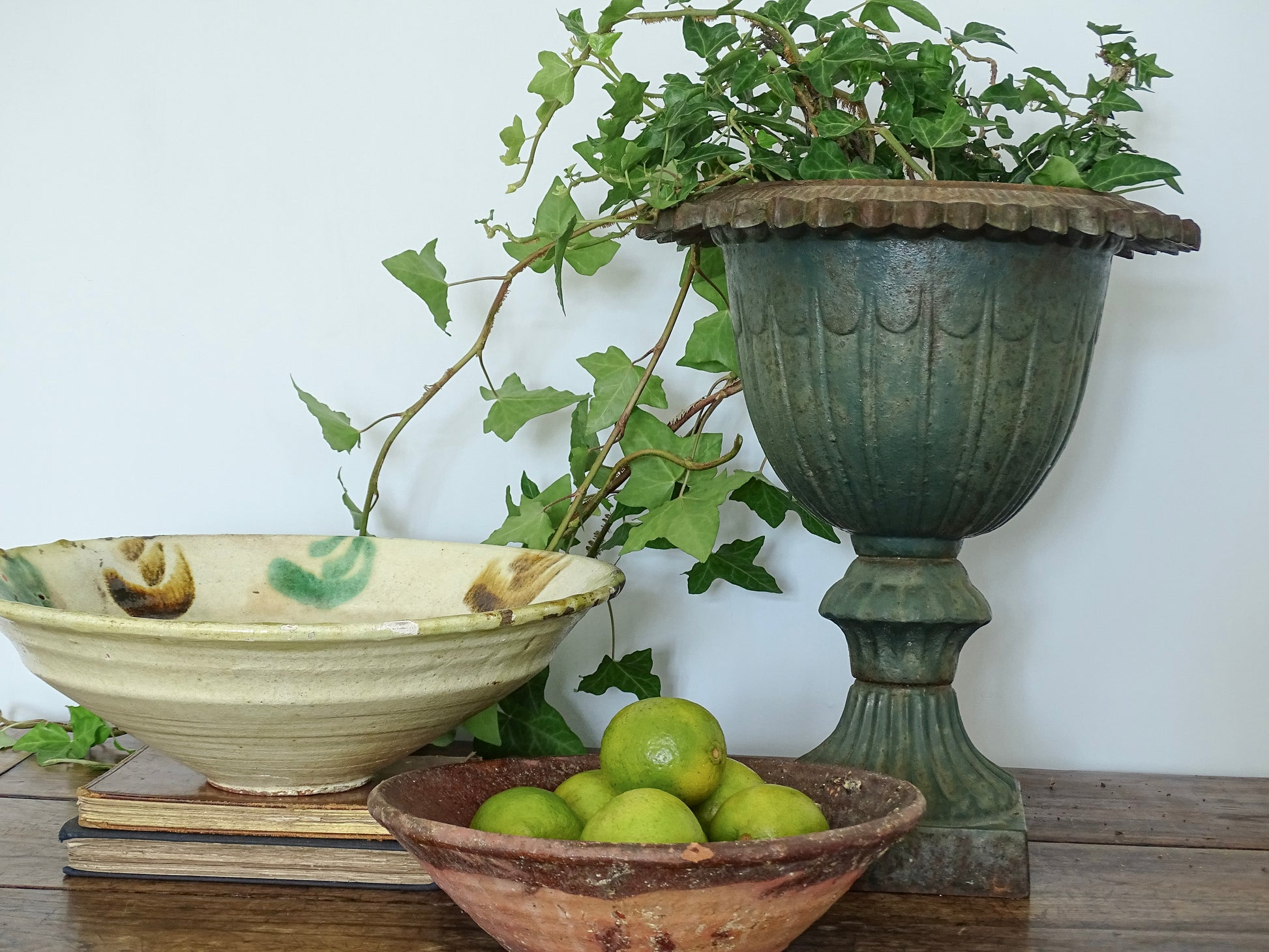 Large French antique cast iron urn planter with green ivy, earthenware bowl with manganese glaze resting on stacked books, and a terracotta bowl filled with green limes on a rustic wooden table against a pale blue wall in a French country house