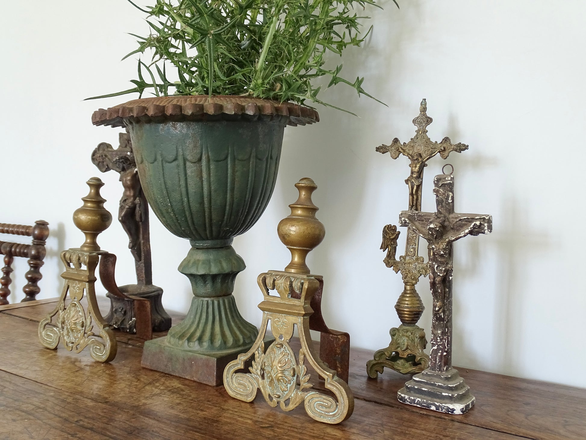 French antique cast iron urn filled with thistles on a sideboard in a French country home alongside brass andirons and religious decor.