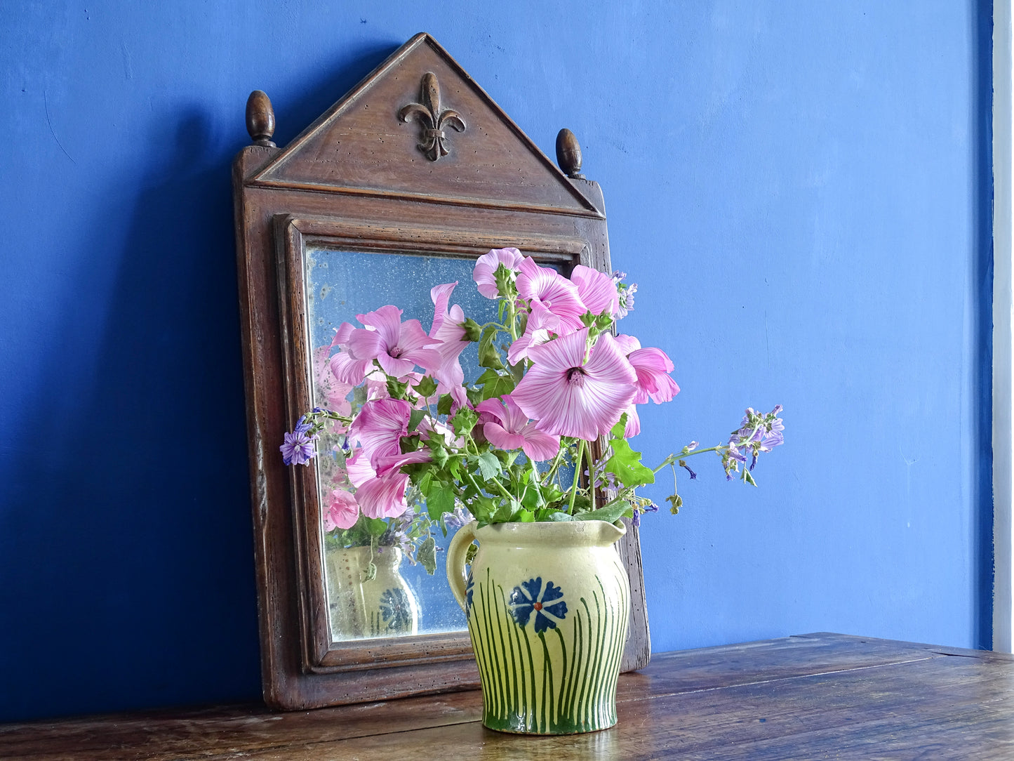 French antique ceramic jug with yellow glaze and hand-painted design filled with pink flowers in front of an antique wooden mirror in a room with painted blue walls