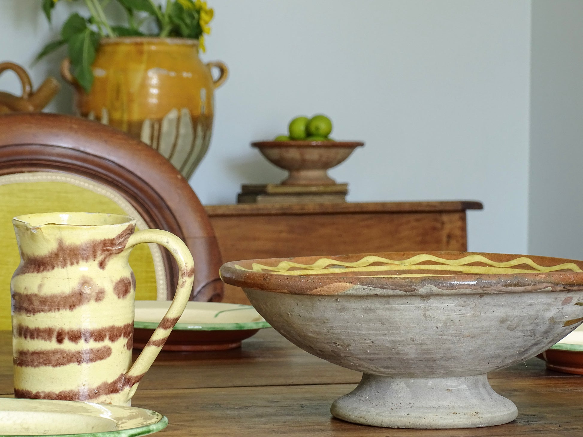 A rustic French antique ceramic pitcher set on a wooden table, with a larger pedestal bowl and a confit pot in the softly blurred background.