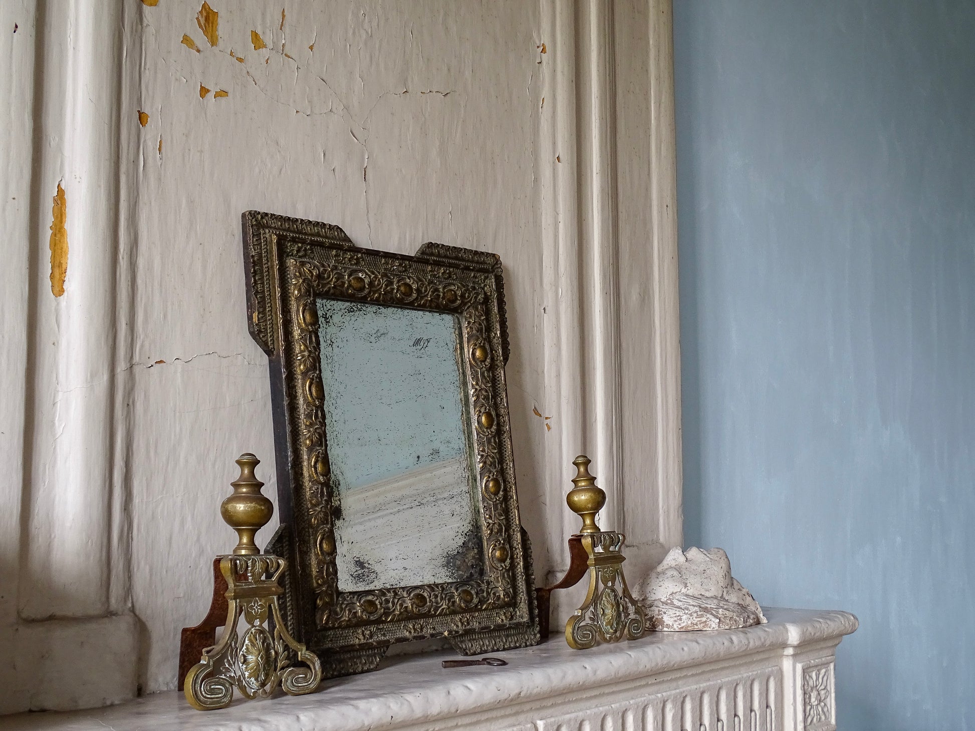 French antique gold mirror with foxed glass on a stone fireplace with brass andirons in a room with pale blue painted walls in a French country house. 