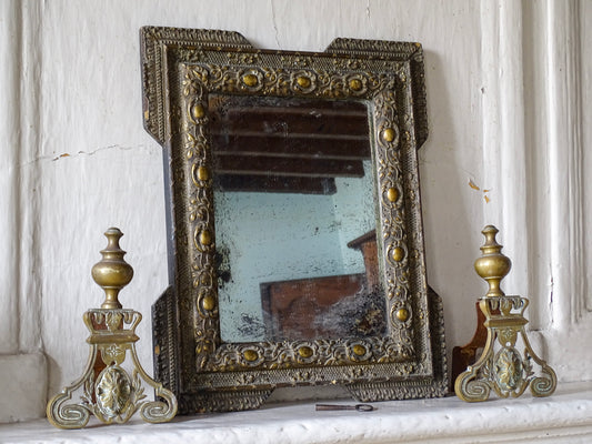French antique gold mirror with foxed glass on a stone fireplace with brass andirons in a room with pale blue painted walls in a French country house. 