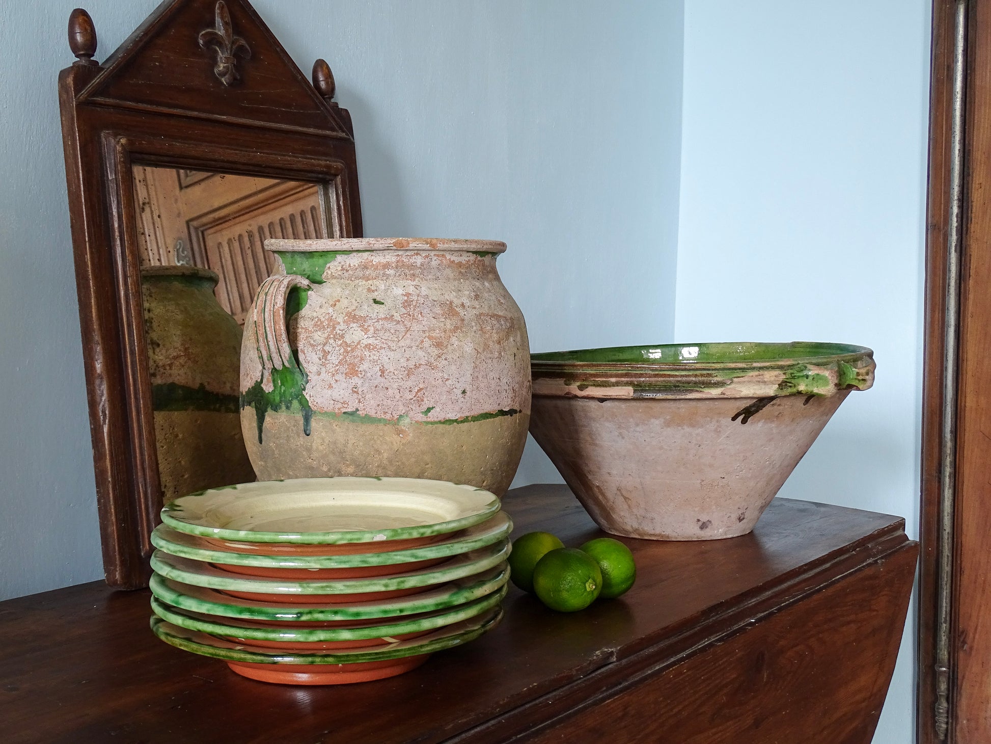 French confit pot with green glazed tian bowl, a stack of earthenware plates and wooden mirror in a French country home with pale blue walls