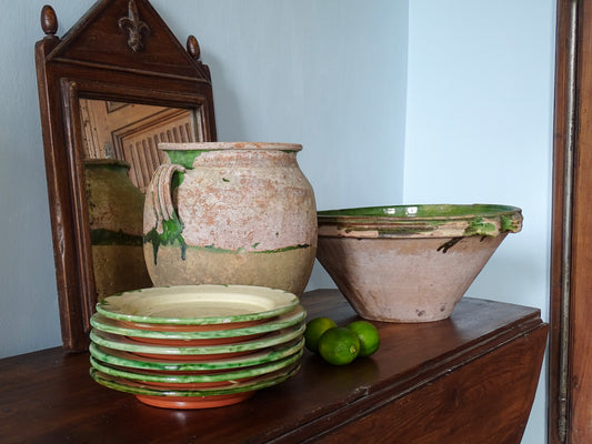 French confit pot with green glazed tian bowl, a stack of earthenware plates and wooden mirror in a French country home with pale blue walls
